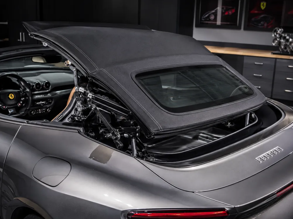 Ferrari grand tourer parked with its roof half-open in a clean workshop, close-up view of the convertible top fabric, stitching, rear window, and hydraulic linkage, aspirational luxury parts photography style